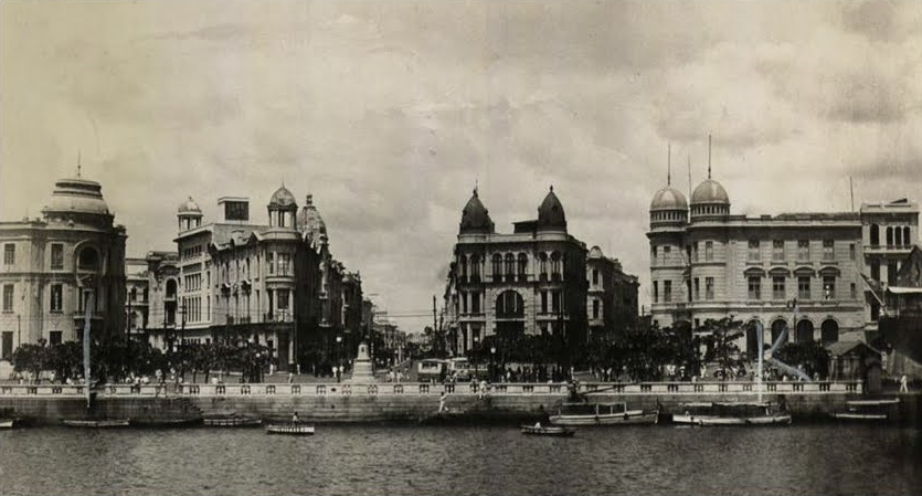 Fotografia em preto e branco do Marco Zero do Recife na década de 1940: casarões com cúpulas à beira do cais e barcos no rio.